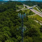 Vista aérea de uma torre de comunicação instalada entre áreas verdes, ao lado de uma rodovia com veículos.