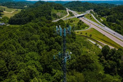 Vista aérea de uma torre de comunicação instalada entre áreas verdes, ao lado de uma rodovia com veículos.