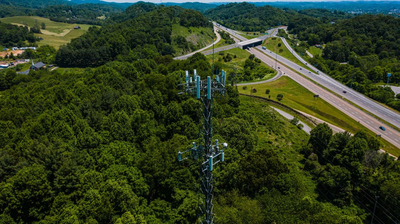 Vista aérea de uma torre de comunicação instalada entre áreas verdes, ao lado de uma rodovia com veículos.