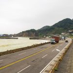 Caminhões trafegam por uma rodovia costeira cercada por montanhas e mar.