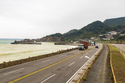 Caminhões trafegam por uma rodovia costeira cercada por montanhas e mar.