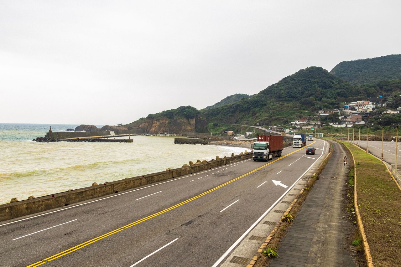 Caminhões trafegam por uma rodovia costeira cercada por montanhas e mar.