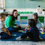 Professora realiza atividade educativa com crianças sentadas no chão de uma sala de aula.